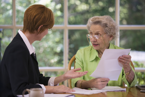 An elderly woman examines a piece of paper, discussing a will-based estate plan with a person in business attire across the table. Sunlight filters through a large window behind them.