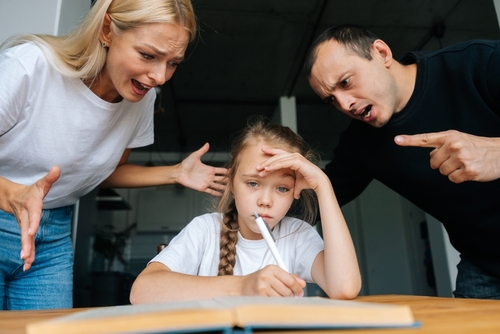 Portrait of angry young parents yelling and scolding together lazy little daughter sitting at table, doing homework, sad looking at camera. Concept of parent disciplining child for bad education.