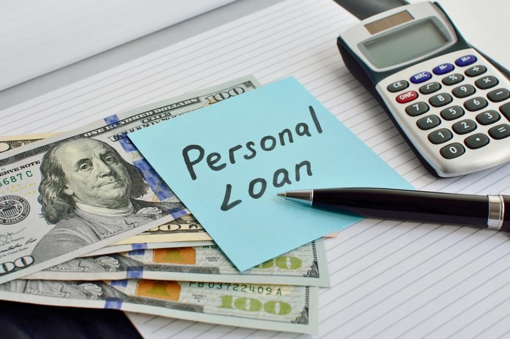 A stack of money sitting on a blank notebook paper with a sticky note attacked to the money saying personal loan and a calculator next to the money 