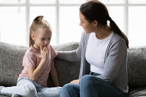 A woman and a child sit on a couch in a bright room. The woman, mindful of what not to say during a custody battle, speaks gently as the child listens with a thoughtful expression.
