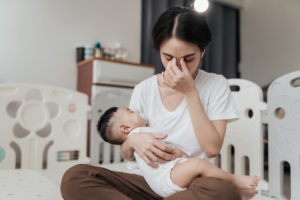A woman sits on the floor holding a baby in her lap, pinching the bridge of her nose with her eyes closed