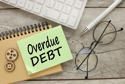 A sticky note with "Overdue Debt" sits atop a notebook, surrounded by gears, glasses, a pen, and a keyboard on a wooden surface—quiet reminders of financial obligations. Ever wonder if a collection agency can sue you after 7 years? It's an important question to consider amidst deadlines.