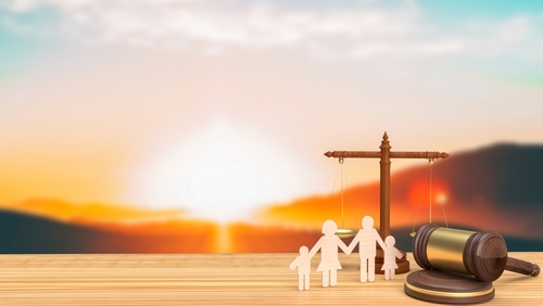 Scale balancing paper family figures and a gavel on a wooden table against the backdrop of a serene sunset.