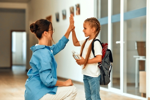 In a hallway, a woman kneels to high-five a young girl with braided hair and a backpack, reminding her of the importance of positivity even during challenging times like custody battles.