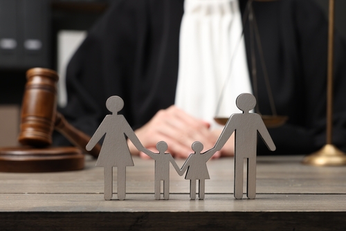 Wooden family cutouts stand on a table before a gavel and scales, subtly hinting at the complexities of custody battles, with a person in judicial attire in the background, perhaps pondering if living with a boyfriend can affect custody arrangements.