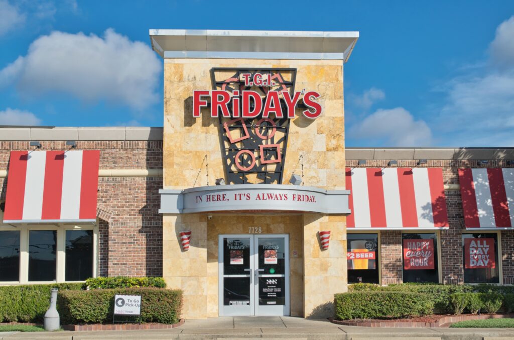 Exterior of a TGI Fridays restaurant with red and white signage, striped awnings, and a entrance featuring the phrase "In Here, It's Always Friday.
