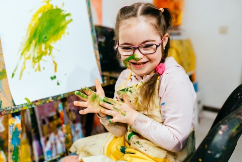 A child with glasses, covered in green and yellow paint, smiles next to a canvas. In a pink shirt and apron, she joyfully engages in painting, creating vivid brush strokes that embody her boundless creativity.