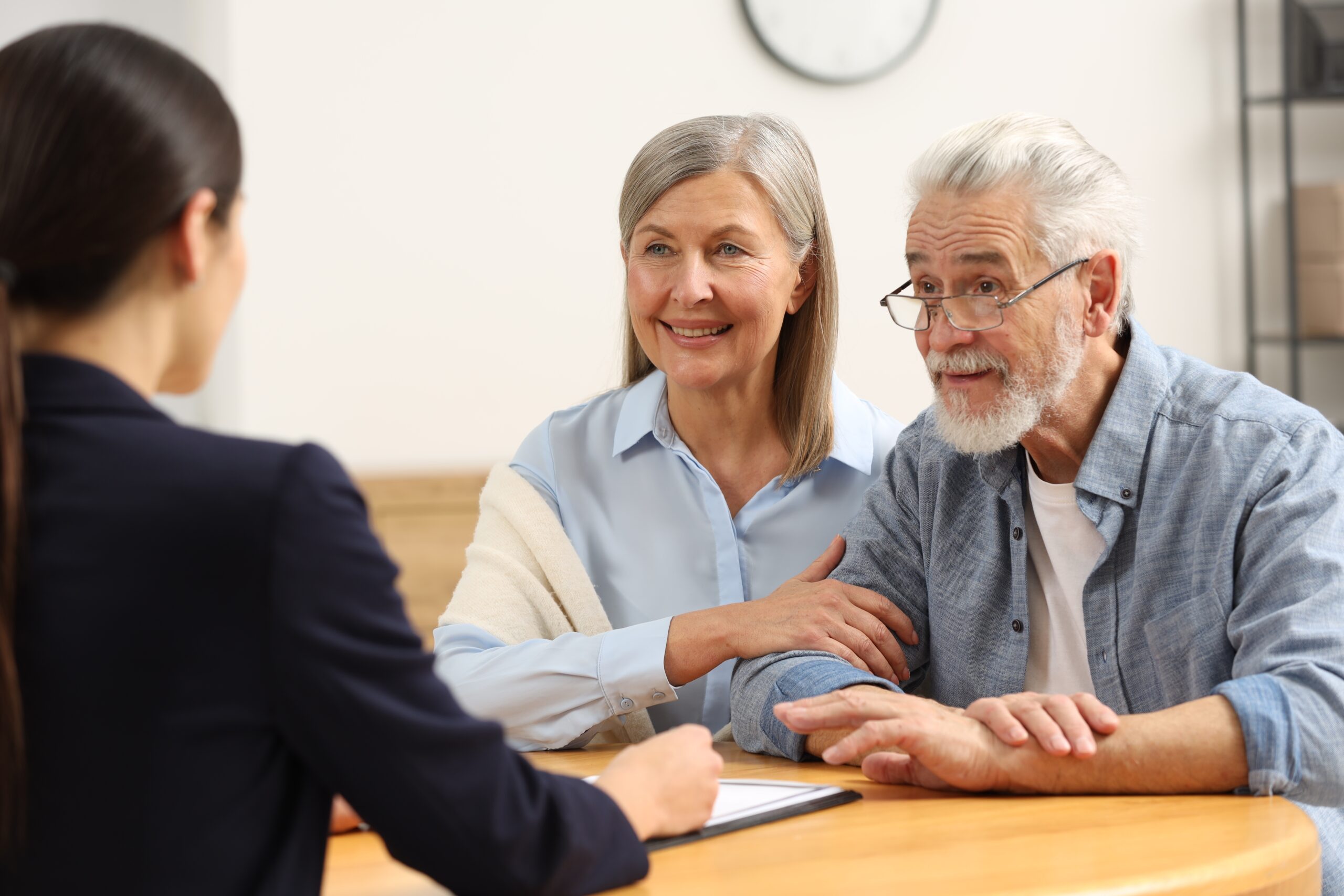 An older couple listens attentively to a professional woman across a table. The woman holds a tablet and gestures while the couple appears engaged in conversation.