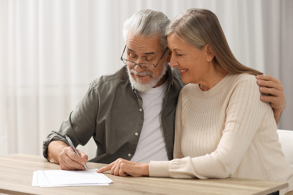An older man and woman sit together at a table, with the man signing a document following will signing instructions while the woman smiles and points at the paper.