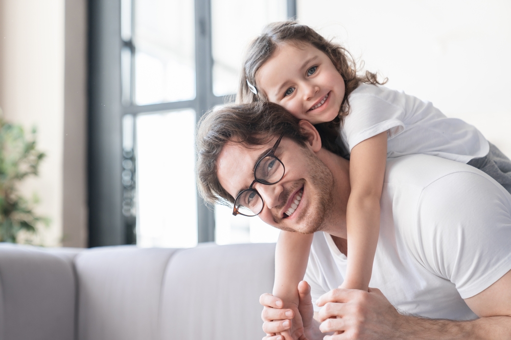 A man with glasses smiles while a young girl hugs him from behind. They are both indoors, sitting on a couch, and looking at the camera—capturing a moment 