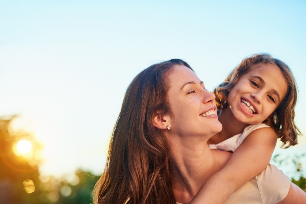 A woman gives a young girl a piggyback ride outdoors at sunset, both smiling happily against a clear sky, enjoying quality time together reminiscent of special moments like Mother's Day under a standard possession order in Texas.