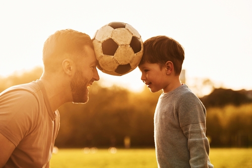 An unmarried father and his child share a joyous moment, smiling as they balance a soccer ball between their foreheads in the fresh Texas air.
