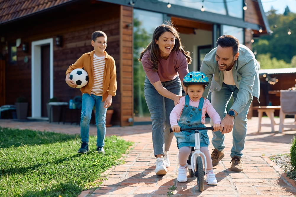 A young girl rides a balance bike outside a house, assisted by two adults—highlighting the benefits of 50/50 custody. Another child stands nearby holding a soccer ball.