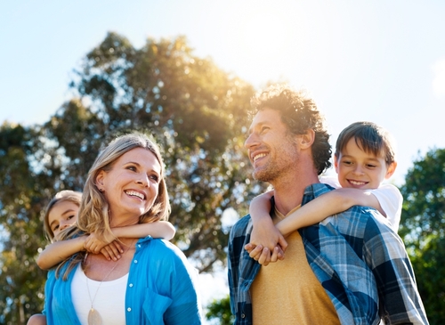 A smiling couple is outdoors with two children on their backs, enjoying the fresh air. They stand in front of the trees under a clear sky, savoring family time without worrying about when child support ends in Texas.
