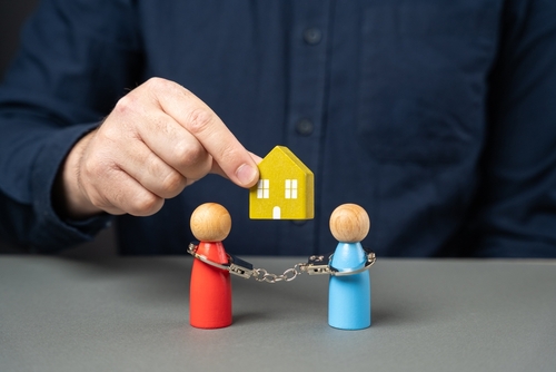 A person holds a small yellow house model above two wooden figures linked by chains, symbolizing shared ownership. This resonates with the complexities of how assets, including those held overseas, are treated in a divorce.
