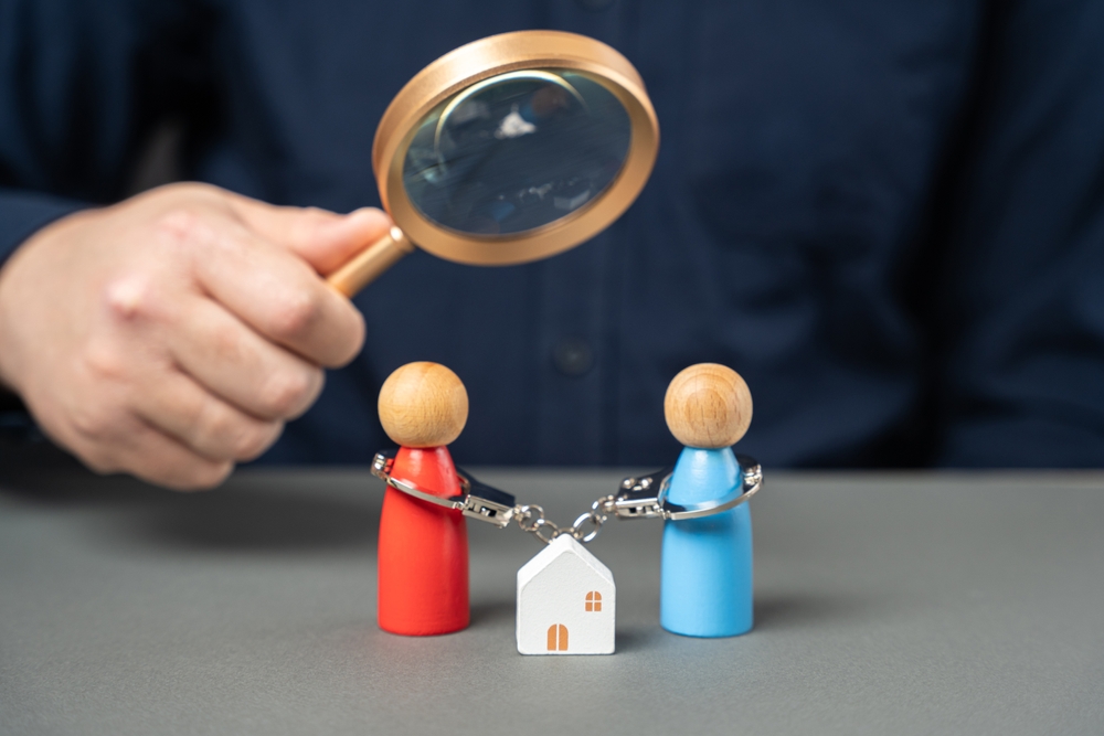 Dividing property after divorce. legal consultation. determine ownership. Divorce proceedings. A man holds a magnifying glass over a handcuffed couple standing near a house.