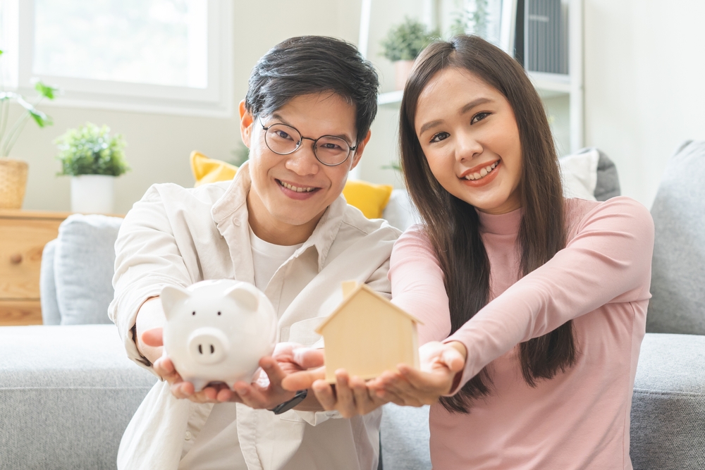 Happy asian family young couple love hand holding piggy bank to save and model house for saving money to buy real estate for new home. Business finance, deposit with banking for financial planning.