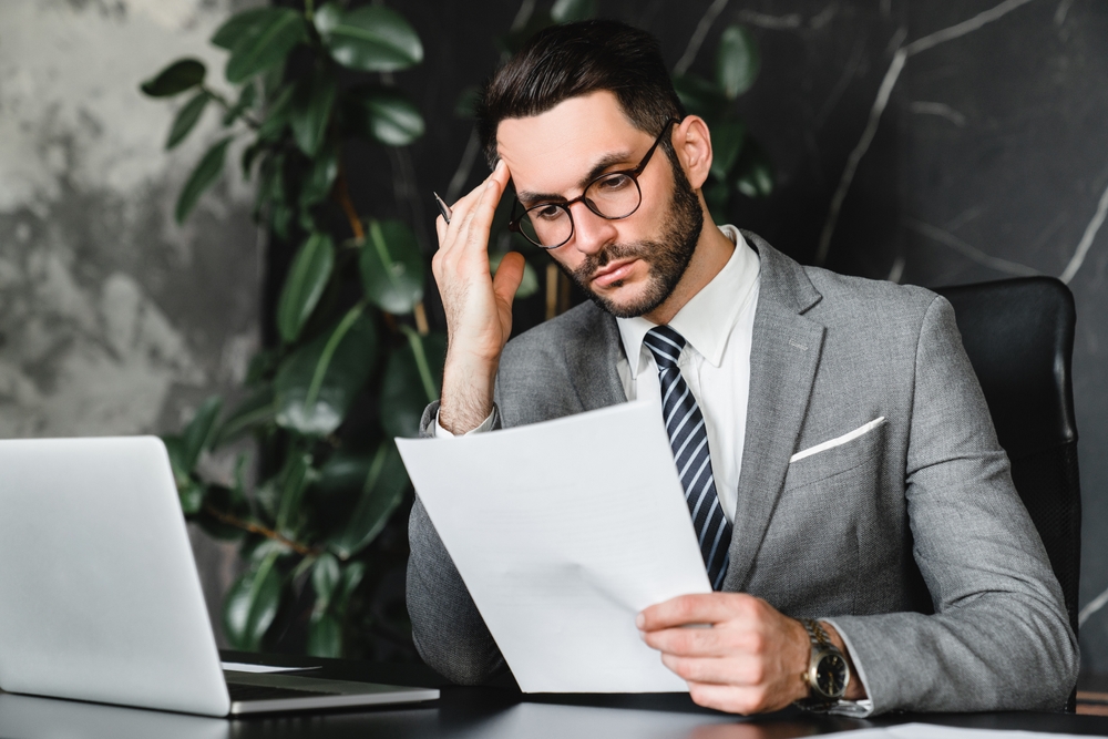 Caucasian stressed businessman checking documents while working on laptop online from office