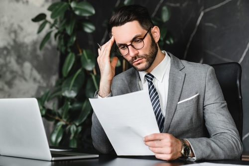 A man in a gray suit and glasses sits at a desk, reviewing what documents he needs to file for bankruptcies while looking at a laptop. A large plant is in the background.
