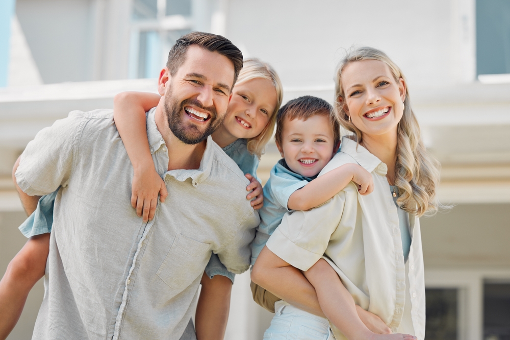 A smiling family of four poses outdoors, with the parents carrying their two young children on their backs in front of a light-colored house.