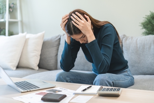 Amidst a sea of papers, a laptop, and a calculator on the couch, a person clutches their head with both hands, overwhelmed by financial stress and pondering if excluding a credit card from Chapter 7 is even an option.