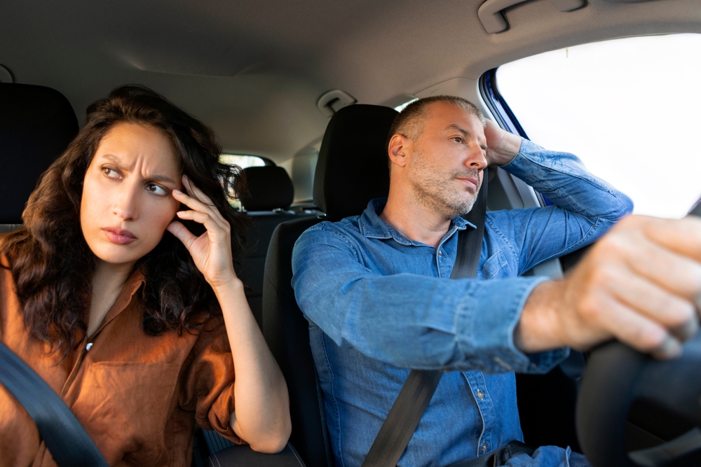 Upset woman sitting next to her husband driving auto, touching head and look aside, feeling down after quarrel with spouse