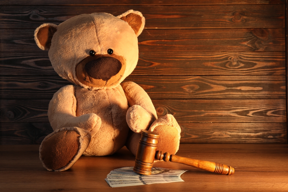 A plush teddy bear sits next to a wooden judge’s gavel and a stack of cash on a wooden surface, symbolizing how to prove you are a fit parent in court, set against a wooden wall background.