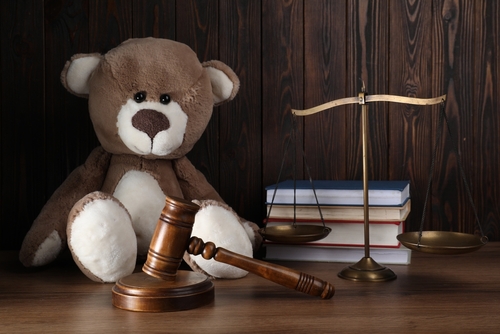 A teddy bear sits next to a gavel and a balance scale on a wooden table, symbolizing justice, while a stack of books on unmarried father rights in Texas looms in the background.