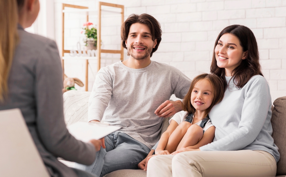 A man, woman, and young girl sit together on a couch, smiling and talking with a person holding a clipboard about the standard possession order in Texas for Mother’s Day in a bright living room.