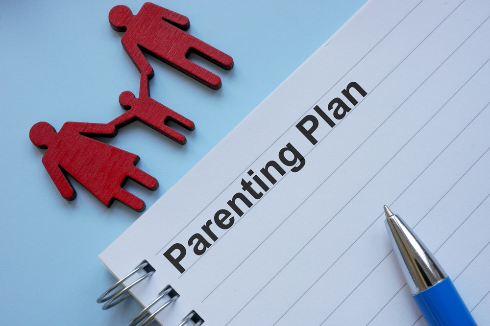 A notepad labeled "Parenting Plan" with a blue pen and red cutout figures of two adults and a child holding hands, illustrating the question: what does full custody mean, on a light blue background.
