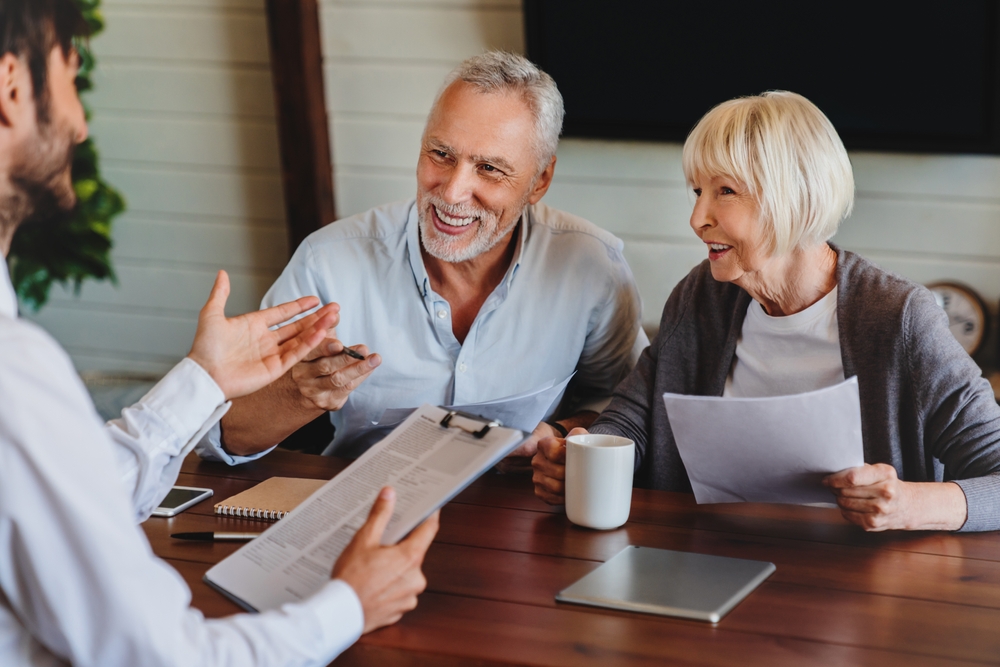 An older couple sits at a table, smiling and holding documents, while discussing will with a person who has a clipboard.