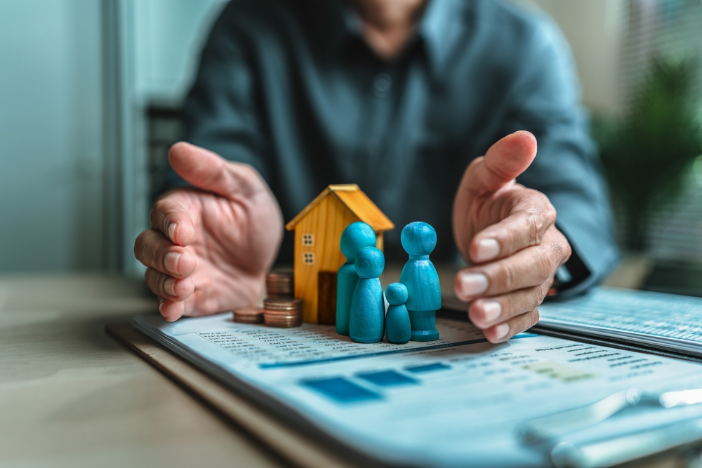 Businessman's hand gestures to protect money, house, wooden dolls represent family. Image represents the concept of asset protection