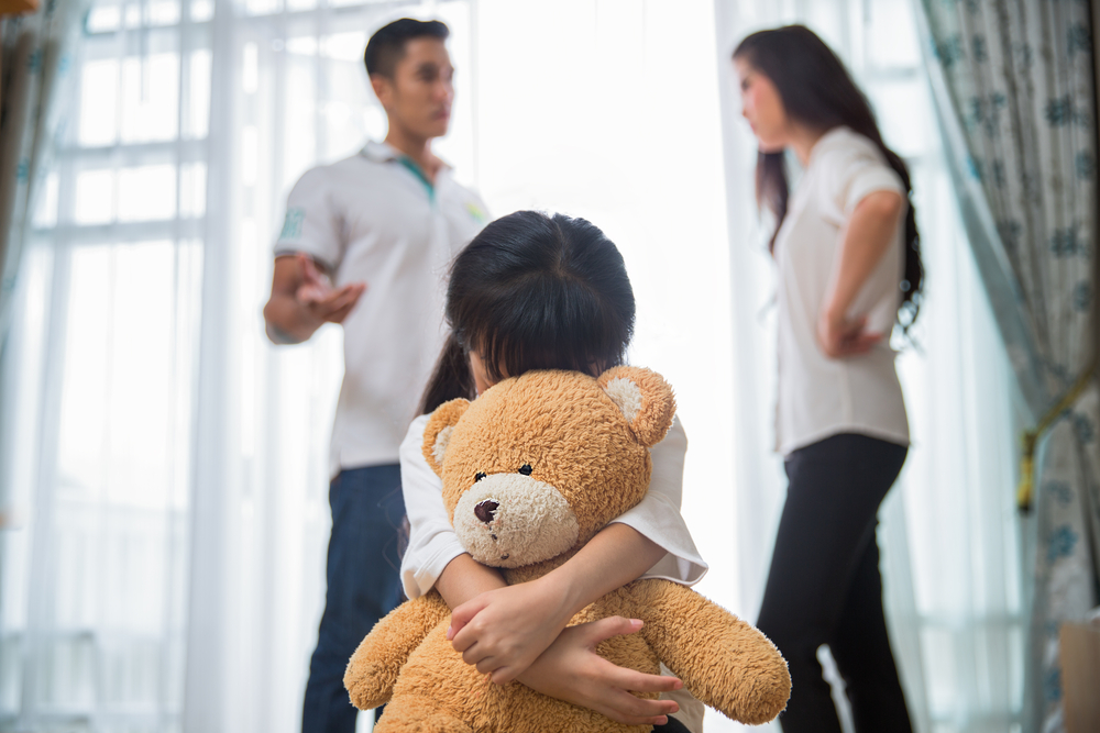 A young girl hugs a teddy bear while two adults argue in the background, hinting at a possible texas unfit parent situation inside a brightly lit room.