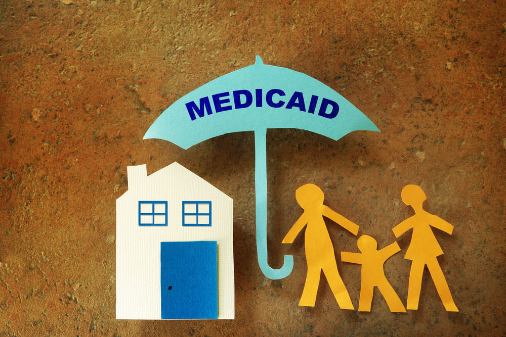 Paper cut outs of a family holding an umbrella that says medicaid over themselves and a paper cut out of their house 