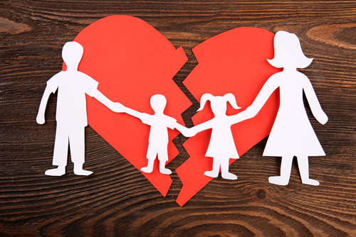 Paper cutouts of a family holding hands stand on a wooden surface, casting shadows around a large, broken red heart symbolizing separation or divorce. This poignant scene reflects the delicate matters like custody in Texas divorces.