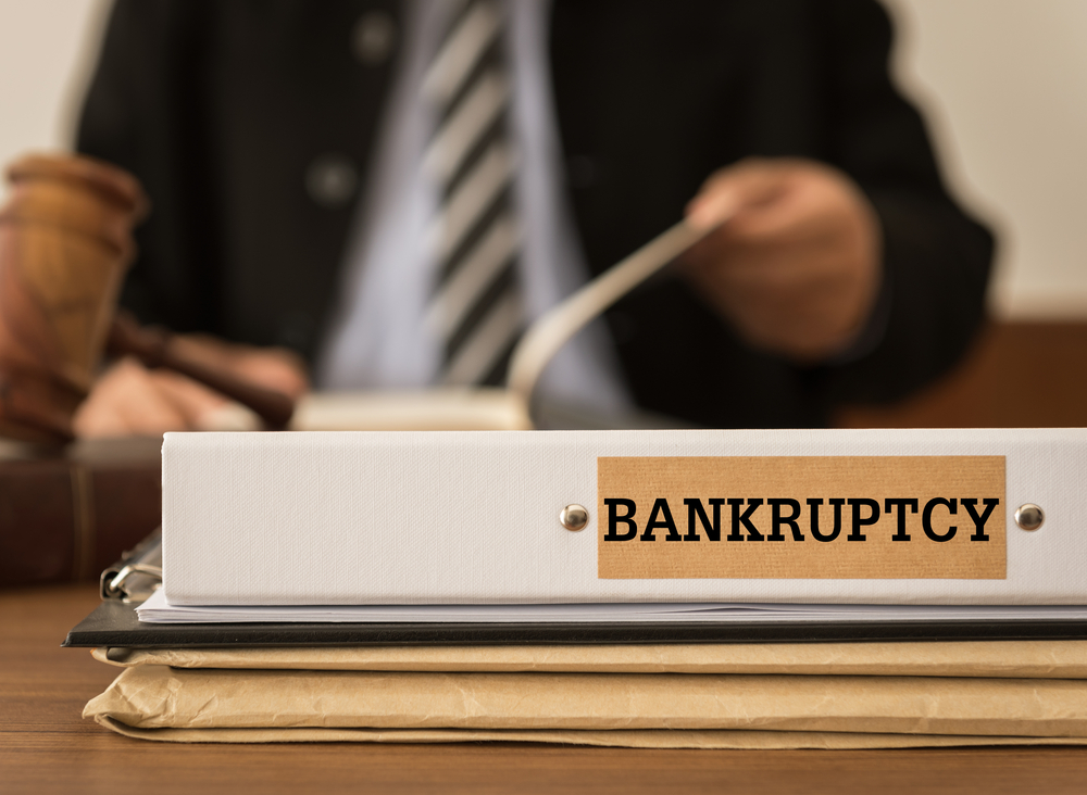 A binder labeled "Bankruptcy" sits on a desk with documents, while a person in a suit and a gavel are visible in the background—raising questions like, can I be sued twice for the same debt?.