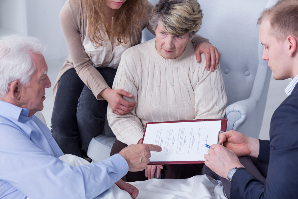 An elderly woman sits in a chair, supported by a younger woman, one pointing and the other holding a pen.