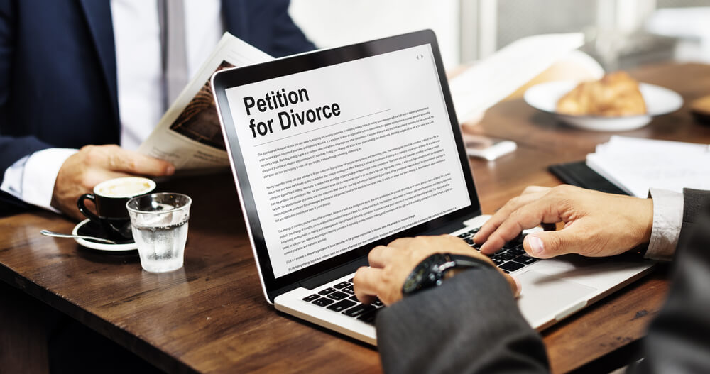 A businessman sitting at a table with his laptop open and a petition for divorce on the screen of his computer