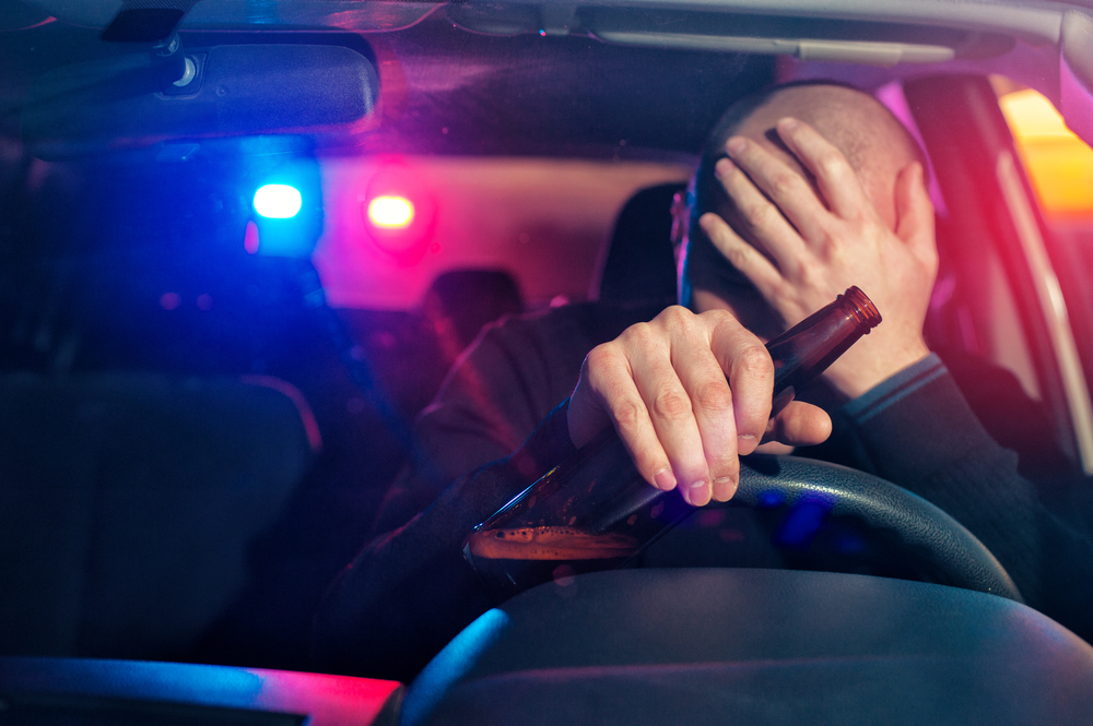 A person sits in a car holding a beer bottle, covering their face with their hand, as police car lights flash in the background