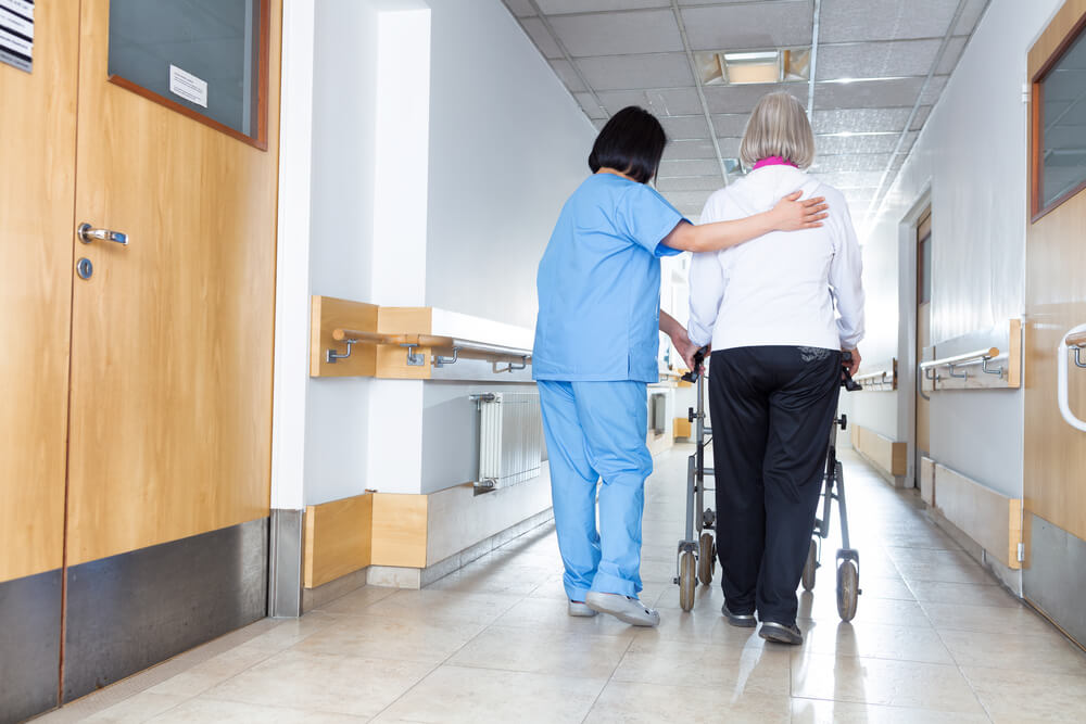 An image of a nurse helping an elder walk down the hall of a nursing home