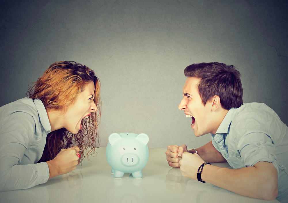 Finances in divorce concept. Wife and husband can not make settlement screaming piggy bank in-between sitting at table looking at each other with hatred