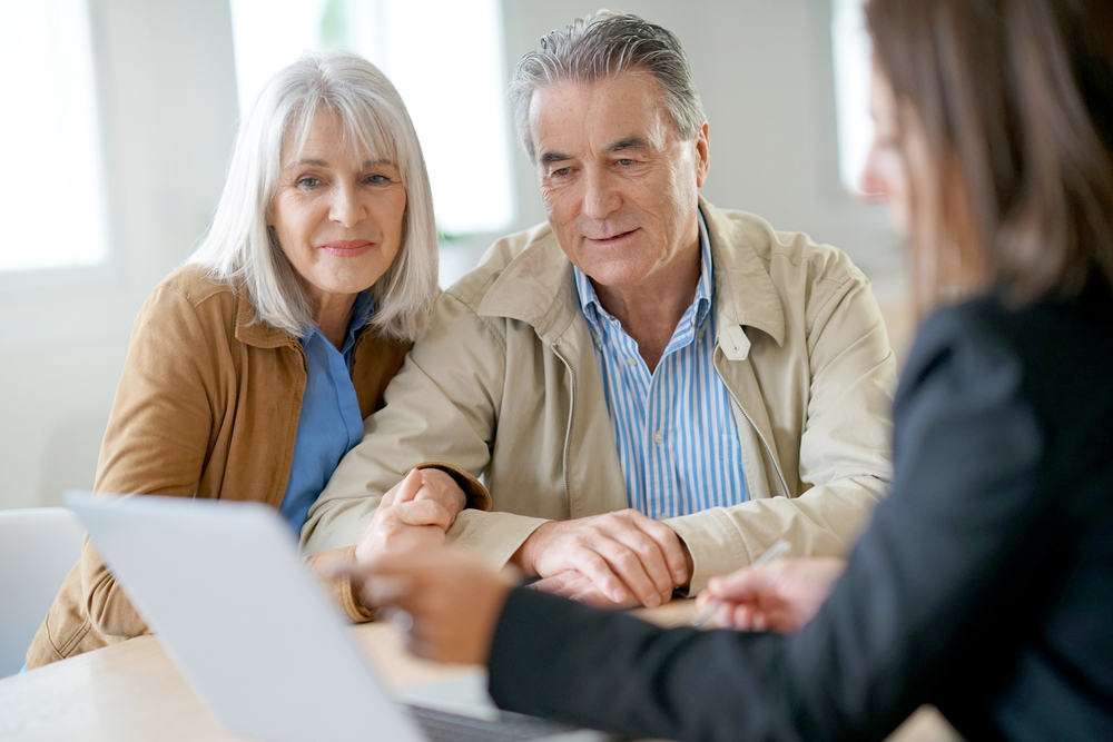 An older couple sits at a desk, listening to a professional woman who is explaining the process of creating a joint will on her laptop.