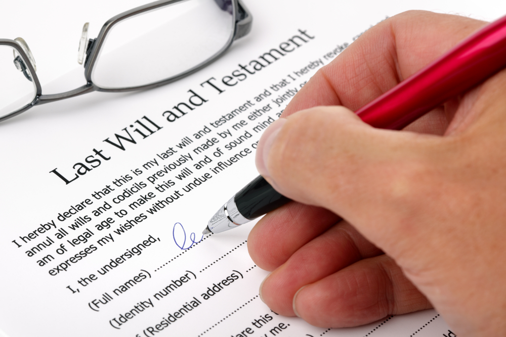 A hand holding a pen signs a last will and testament form, following will signing instructions, with a pair of eyeglasses resting on the document.