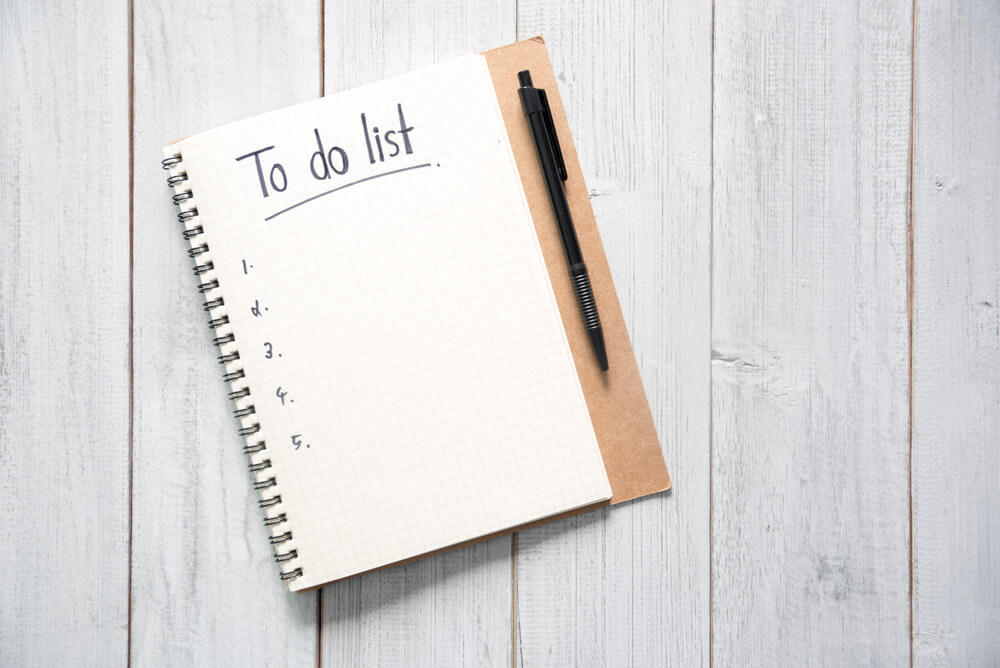 A notebook that is open and on the paper it says to do list with 1-5 listed down the page, a pen is next to the notebook and they are sitting on a wooden table