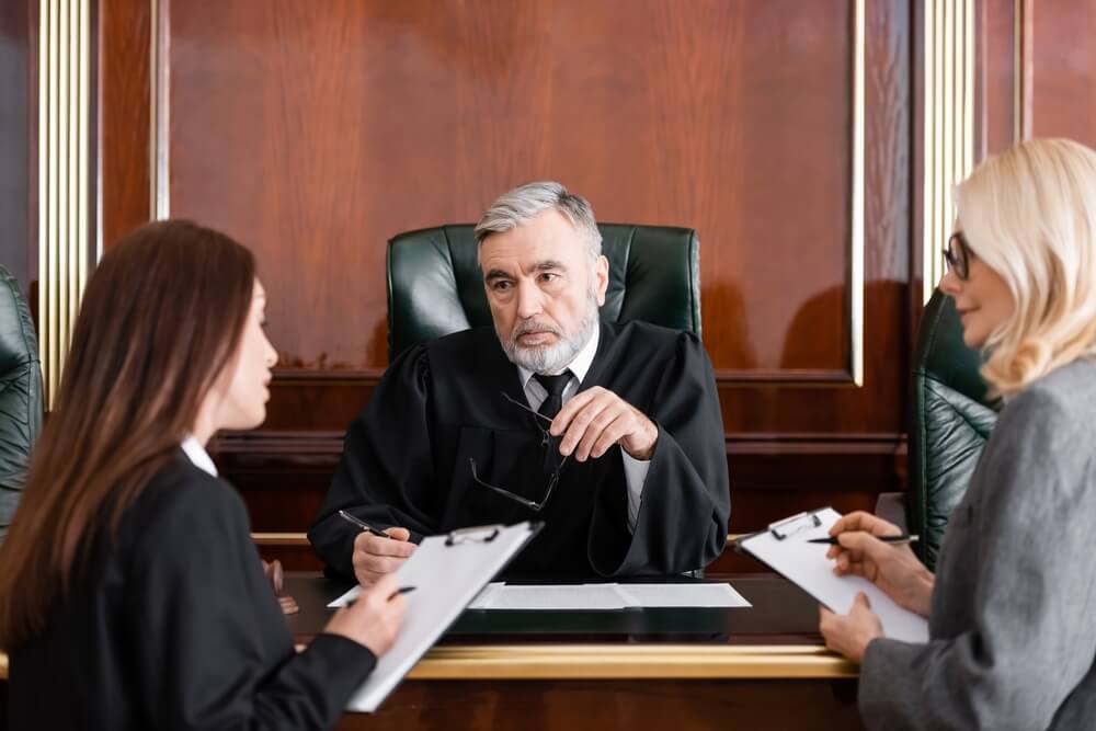 Judge sitting down behind a desk in the middle of two female lawyers holding papers 