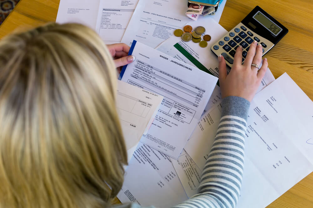 A woman sitting at a wooden table with bank papers spread across the table and the woman is typing into a calculator 