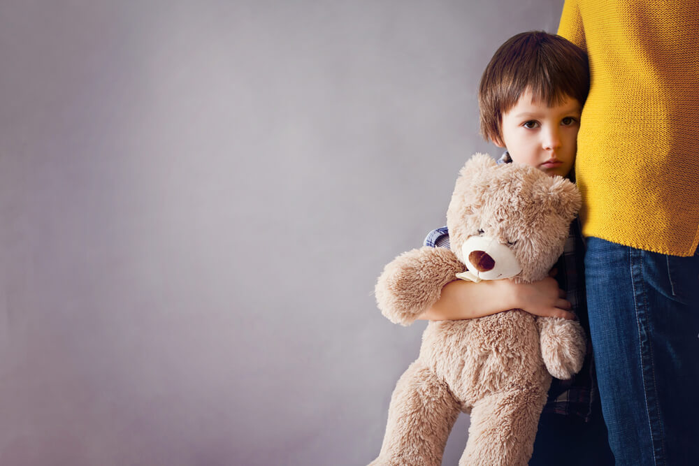 An upset little boy holding a teddy bear leaning on his mothers side 