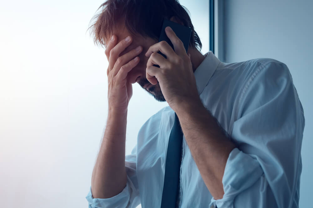 Man on the phone with one hand and his other hand is over his face, he looks frustrated and upset