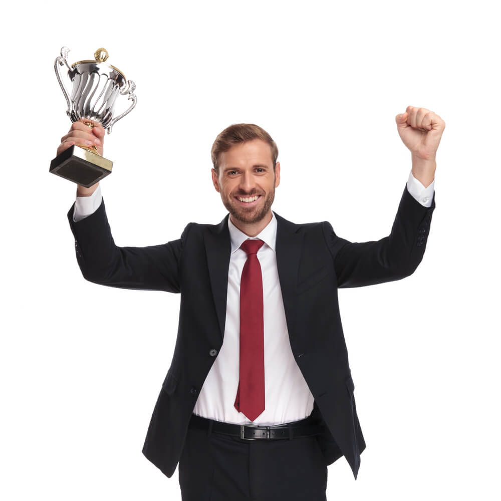 A businessman standing against a white background holding up a trophy in one hand and his other hand in the air celebrating