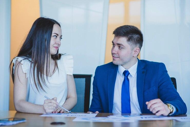 Two people sit at a table in a professional setting, both looking at each other while one writes on papers.